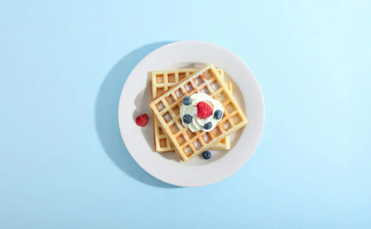 Zwei goldbraune Bananenwaffeln ohne Zucker, garniert mit Sahne, Himbeeren und Blaubeeren, auf einem Teller.
