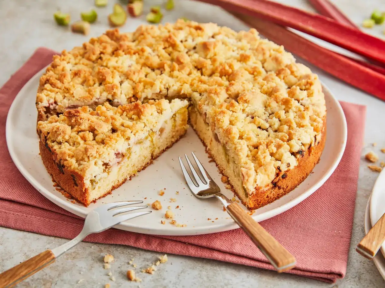 Ein saftiger Rhabarberkuchen mit Streuseln, bereit zum Genießen. Frische Rhabarberstangen liegen daneben.