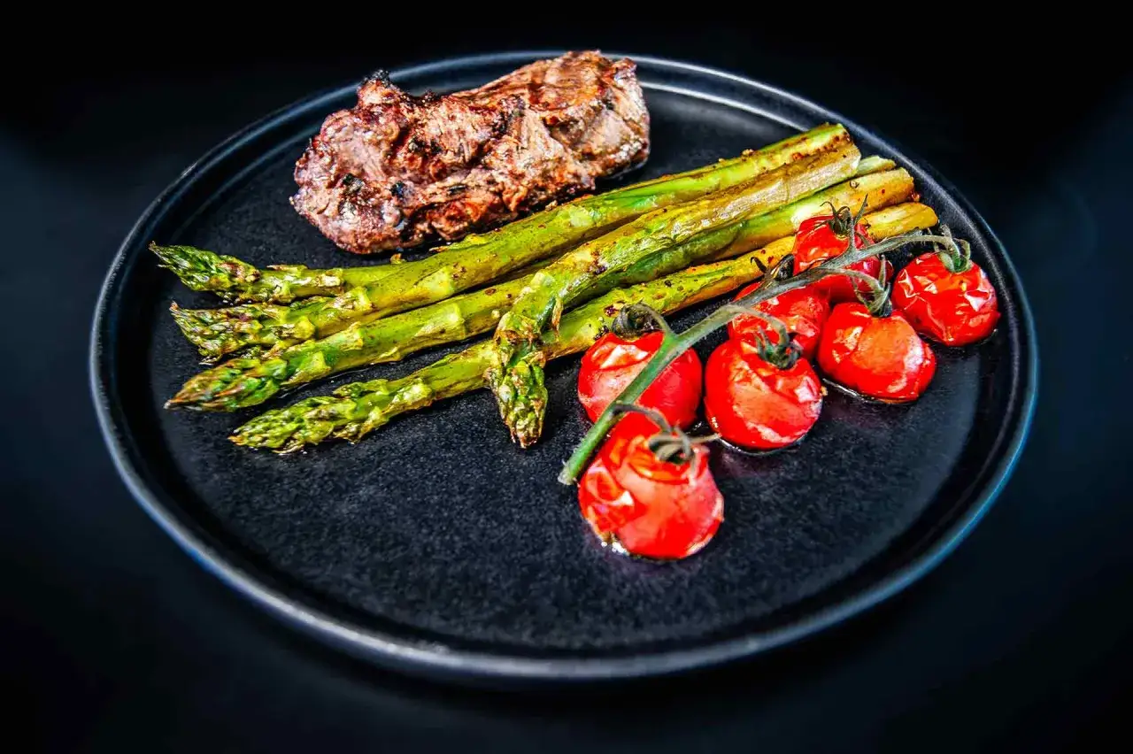 Ein gegrilltes Steak mit gr&uuml;nem Spargel und Kirschtomaten auf einem schwarzen Teller. Ein klassisches Gericht, das zeigt, welches Fleisch gut zu Spargel passt.