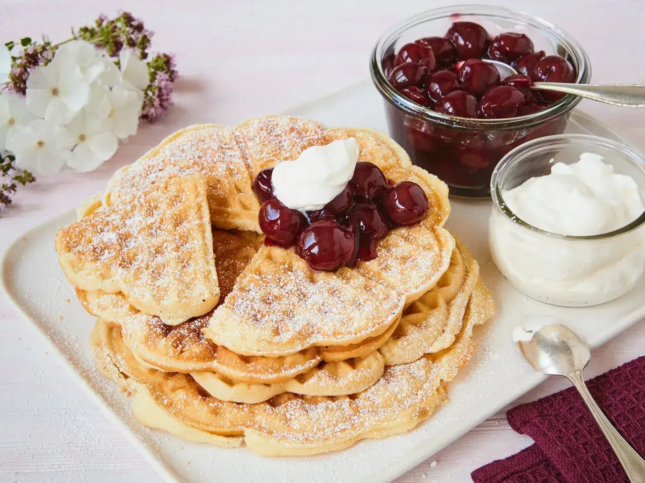 Ein Stapel goldbrauner Waffeln, bestäubt mit Puderzucker, garniert mit Kirschen und Sahne. Ein köstliches Waffeln Rezept für jeden Anlass.