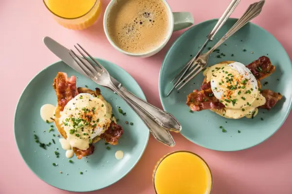 Zwei Teller mit Eggs Benedict, dazu Kaffee und Orangensaft auf rosa Untergrund.